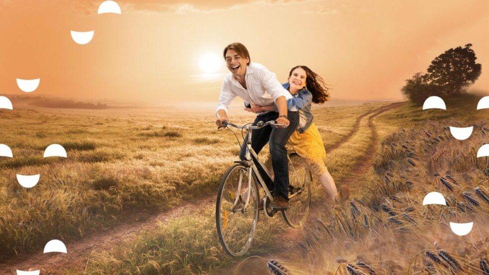 Pariskunta pyöräilee peltomaisemissa - Niin kuin taivaassa kesäteatteri näytelmän mainoskuva