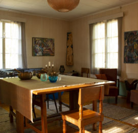 Olo Tila puinen antiikkinen pöytä keskellä huonetta. Seinällä Mikki Paajasen maalaamia tauluja