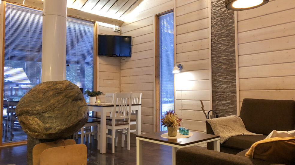 Kaisla- ja Kaarna-mökkien olohuone. Kuvassa näkyy televisio, ruokapöytä sekä kiveen rakennettu takka takaapäin.