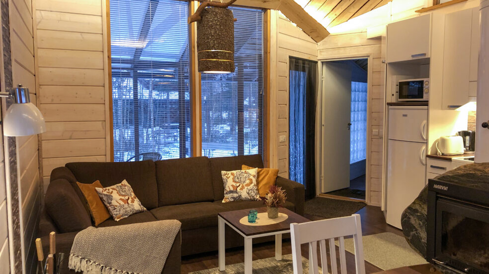 Kaisla- ja Kaarna-mökkien olohuone. Kuvassa on kulmasohva, osa keittiötä sekä kiveen rakennettu takka oikealla.