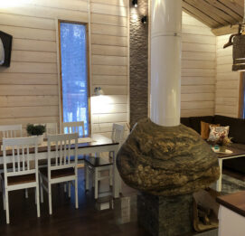 Kaisla- ja Kaarna-mökkien olohuone. Kuvassa näkyy ruokapöytä, kulmasohva sekä kiveen rakennettu takka takaapäin.