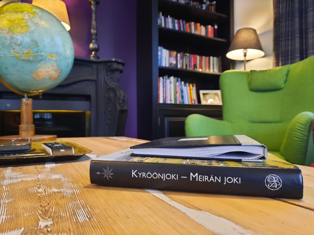Kyröönjoki - Meirän joki kirja Hiljahelenan oleskelutilan pöydällä, vihreä nojatuoli ja kirjahylly taustalla