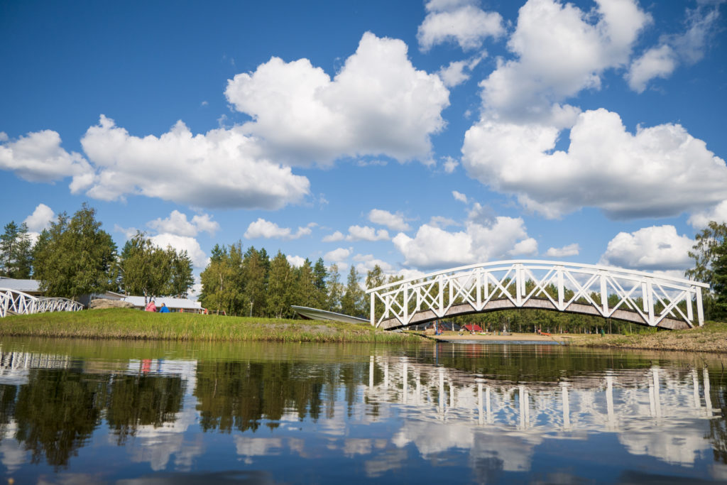 Kalajärven puinen kaareva silta heijastaa tyynessä järvessä sinistä taivasta