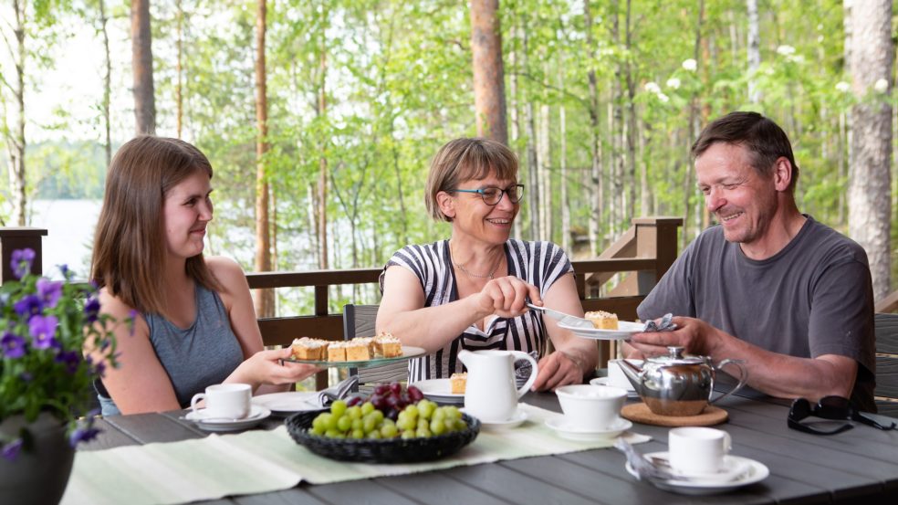 Perhe nauttii aamupalasta mökin terassilla. Metsä ja järvi taustalla.