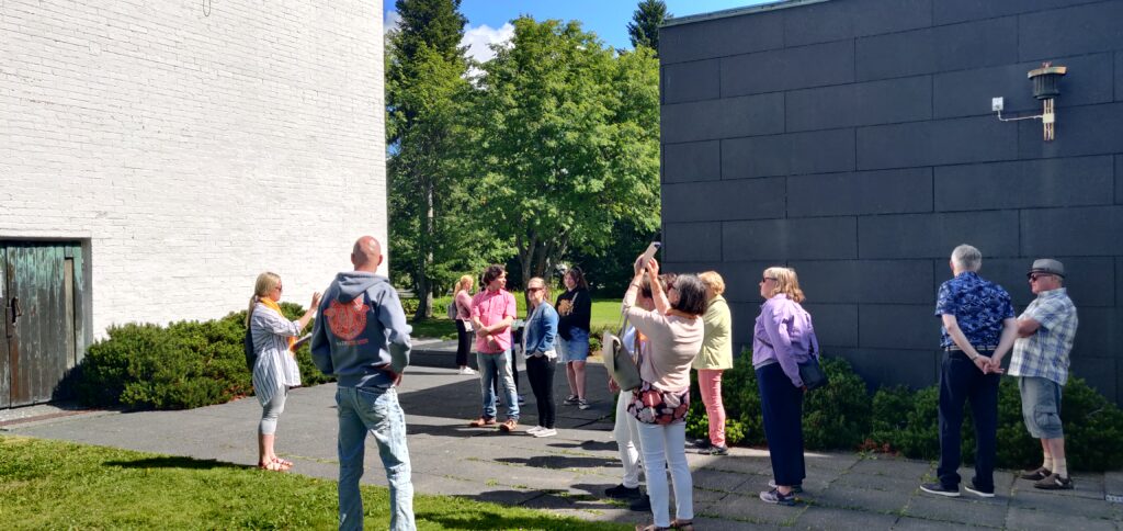 Ryhmä ihmisiä oppaan kanssa Aalto-keskuksessa Lakeuden Ristin tornin juurella ottamassa kuvia aurinkoisessa säässä.